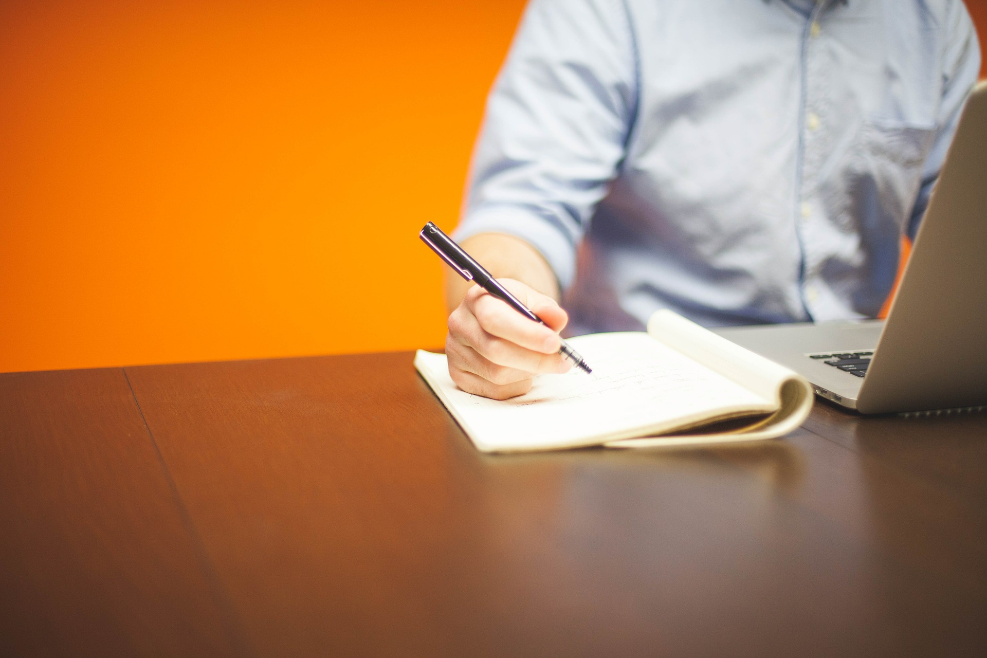 A person writes notes on a notepad whilst reading something off a laptop in a brightly painted room