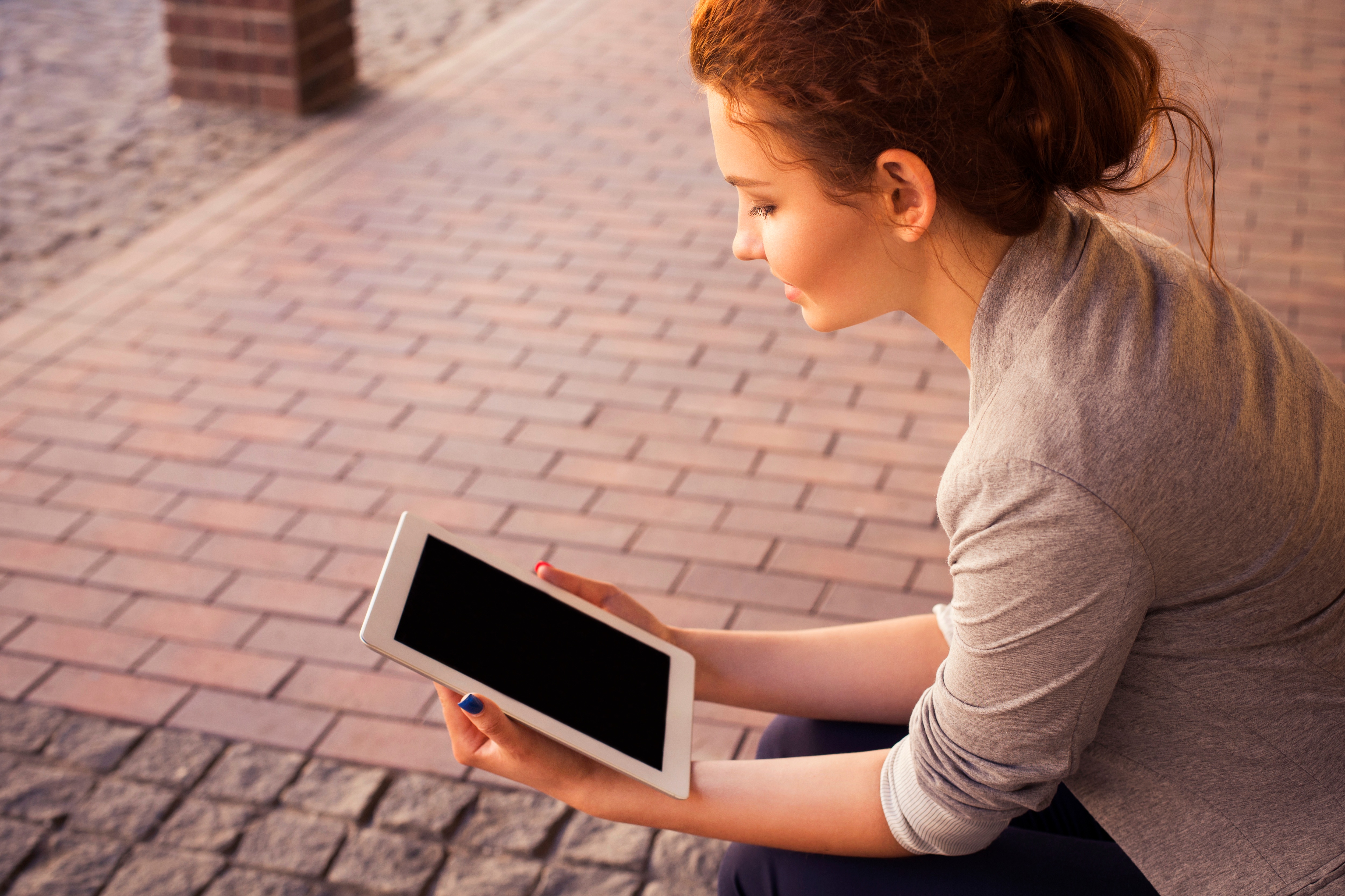 A young lady reads her tablet whilst sat on a brick road