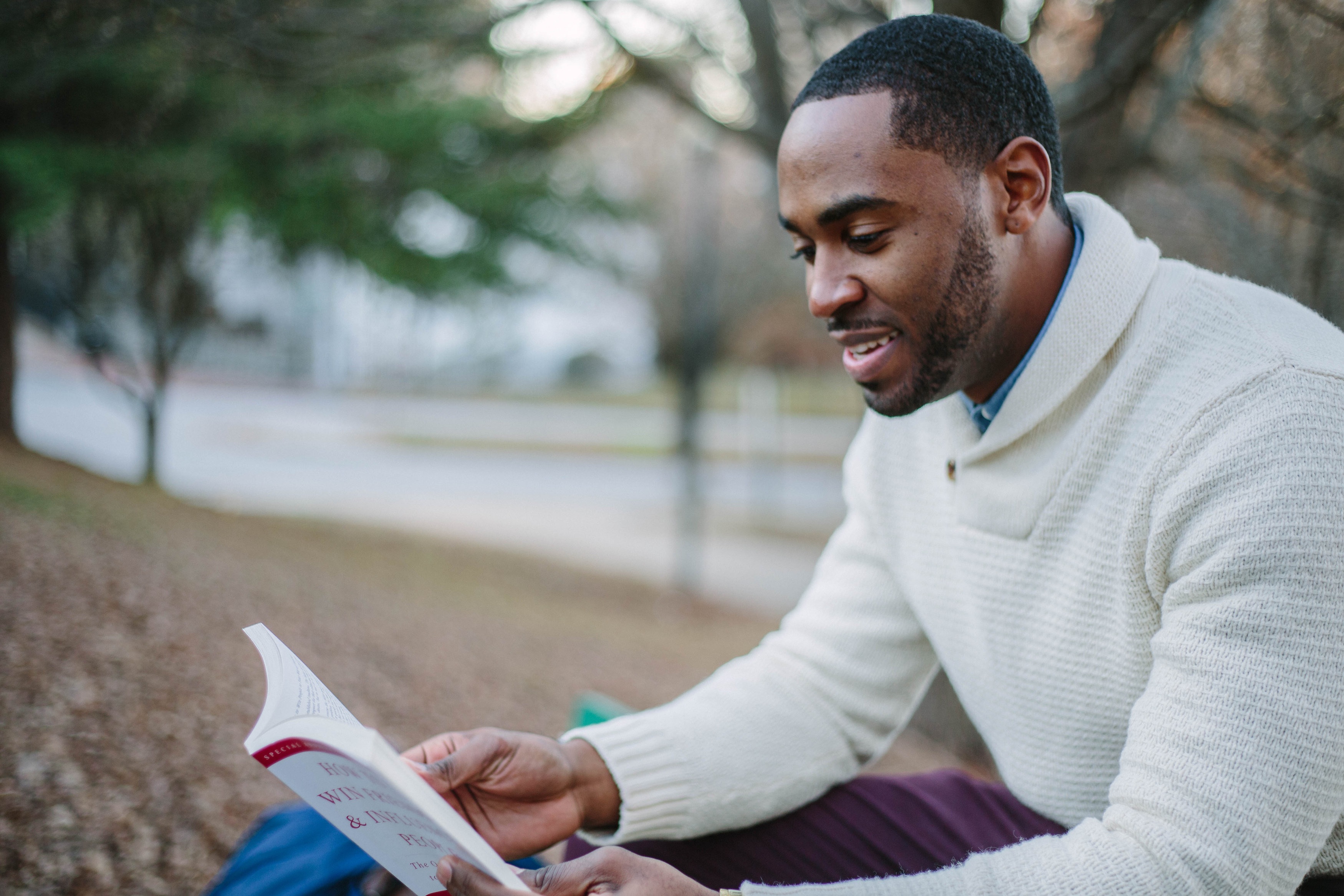 A young man in a thick jumper reading a book in an autumnal park